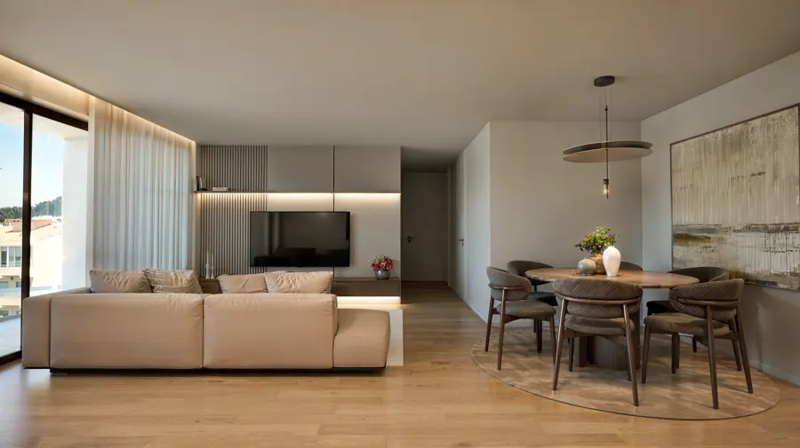 Sala de estar ampla com sofá em tons de areia, pavimento em madeira e cozinha aberta